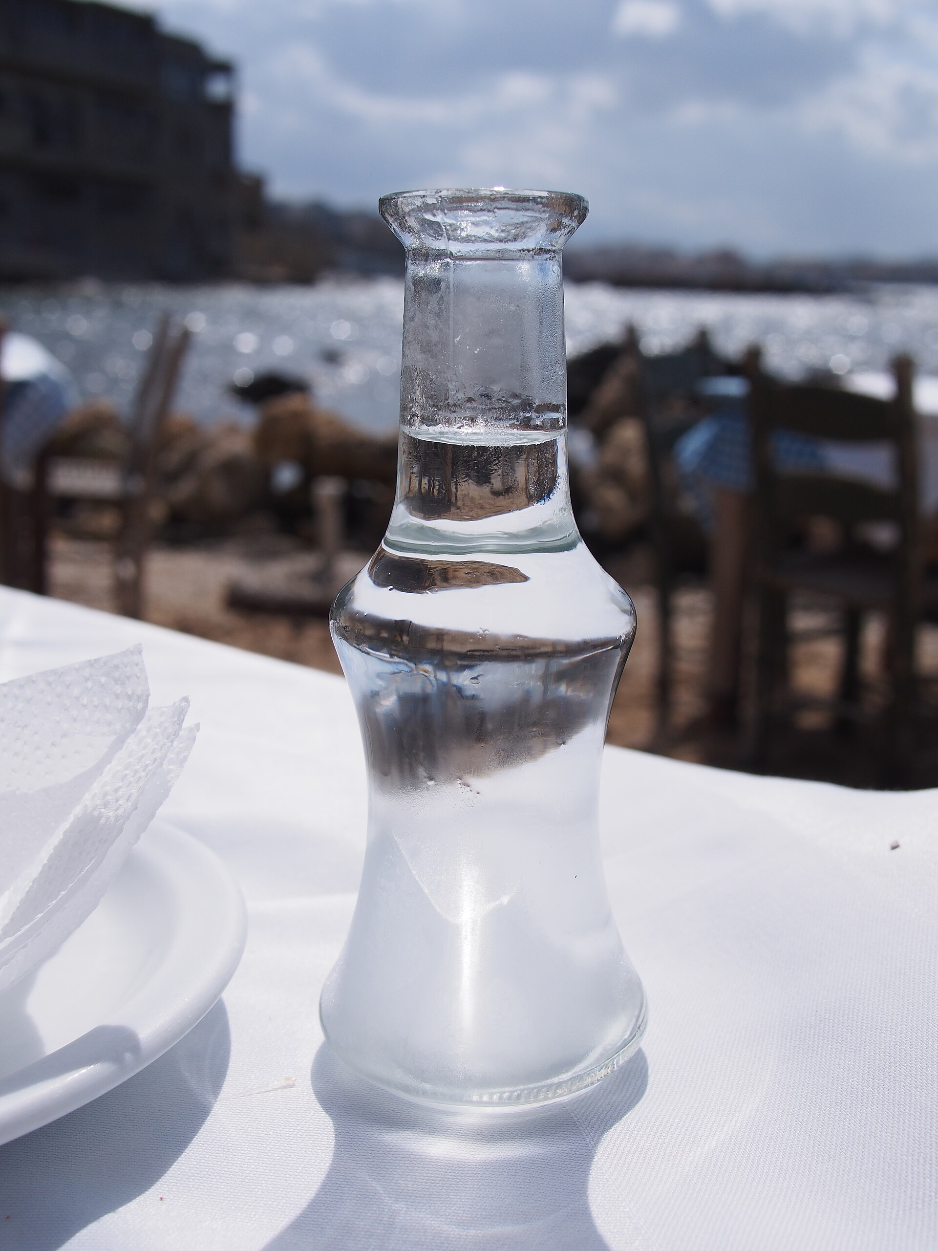 A carafe of homemade tsikoudia (Cretan raki) on a white-clothed taverna table by the waterfront in Chania, Crete, with the harbour behind.