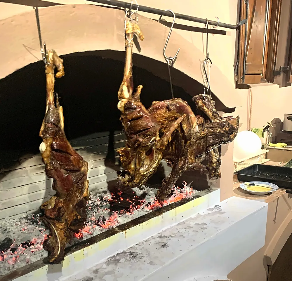 Big cuts of lamb hung on hooks from an iron bar over a bed of glowing embers in a whitewashed village hearth on Crete at Easter