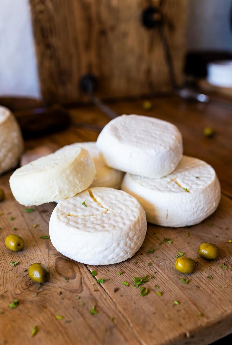Several small rounds of soft white mizithra cheese stacked on a wooden board with green olives and chopped herbs
